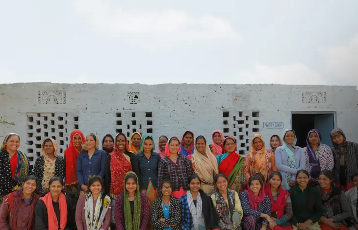 A group of women standing together outside a building with a sign that reads 'WORKSHOP.' They are smiling and wearing various colorful outfits, showcasing a community of empowered women working together.